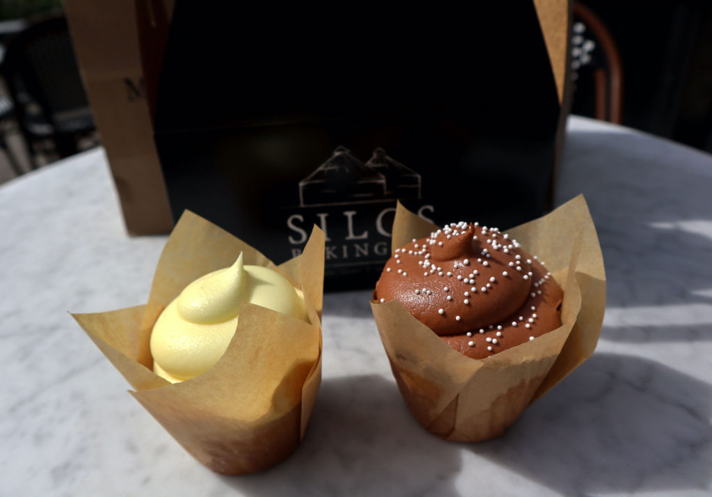 A picture of our two Silo Bakery Co cupcakes next to each other on the outdoor table. One is a yellow lemon frosted cupcake, the other is chocolate frosted with white sprinkles.
